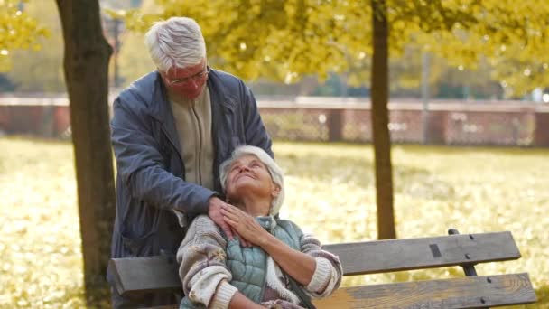 Älteres Ehepaar im Park. Mann legt Hand über die Schulter einer sitzenden Frau - Filmmaterial, Video