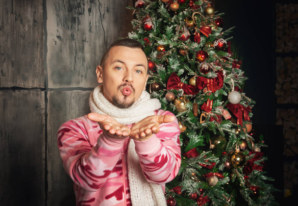 Cheerful funny happy man in pink sleepwear stand near decorated fir tree and sending kiss - Photo, Image