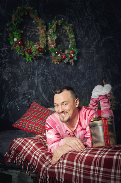 Joyeux homme drôle en vêtements de nuit roses avec cadeau de Noël couché sur le lit - Photo, image