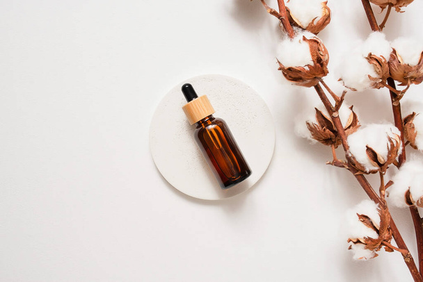 Amber glass dropper bottle with bamboo lid and cotton flowers on white concrete podium. Skincare cosmetic. Beauty concept for face body care. Presentation with daylight. - Фото, зображення