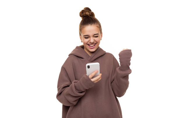 Photo of a beautiful positive cute girl with dark hair and a bun in a brown hoodie with a hood holding a mobile phone, smiling and celebrating a victory isolated on white background with free space - Photo, Image