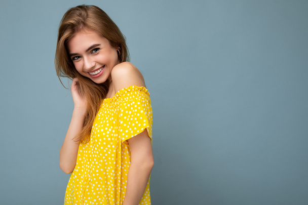 Φωτογραφία του νεαρού χαριτωμένο ευχάριστο όμορφη ελκυστική σκούρα ξανθιά γυναίκα με ειλικρινή συναισθήματα που απομονώνονται σε μπλε φόντο τοίχο με αντίγραφο χώρο για κείμενο φορώντας κομψό κίτρινο φόρεμα. Θετική έννοια - Φωτογραφία, εικόνα