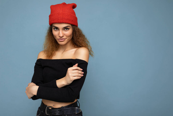 Photo of beautiful self-confident sexy young brunette curly woman isolated over blue background wall wearing black crop top and red hat looking at camera - Valokuva, kuva