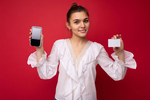 Portrait of positive cheerful fashionable woman in formalwear holding and showing mobile phone with empty screen for mock up and credit card with cut out looking at camera isolated on red background - Fotoğraf, Görsel