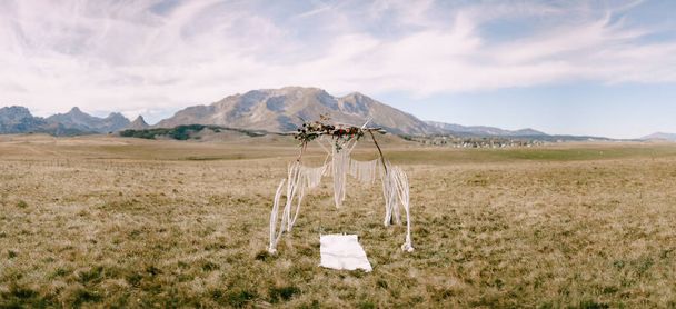 Kırmızı çiçekler ve makrome ile süslenmiş düğün kemeri dağların arka planında bir tarlada duruyor. - Fotoğraf, Görsel