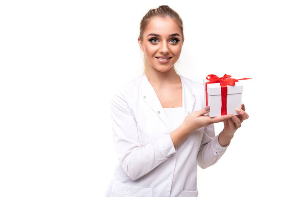 Foto de una hermosa y atractiva joven rubia sonriente positiva con una cola de caballo y maquillaje en un abrigo médico blanco de una enfermera sosteniendo una caja de regalo blanca con una cinta roja aislada en un blanco - Foto, imagen