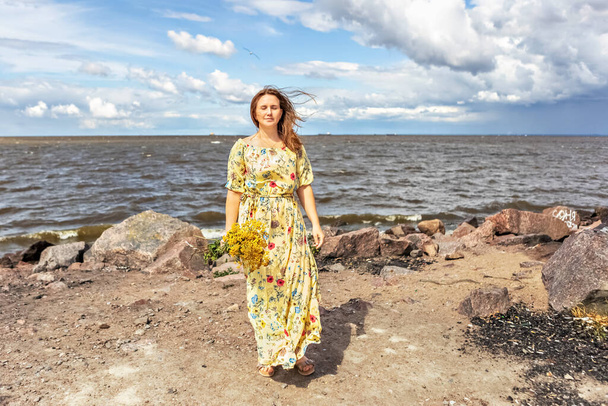 Mladá žena ve žlutých šatech s kyticí tansy stojí ve větru na pláži. Lehké a vzdušné. - Fotografie, Obrázek