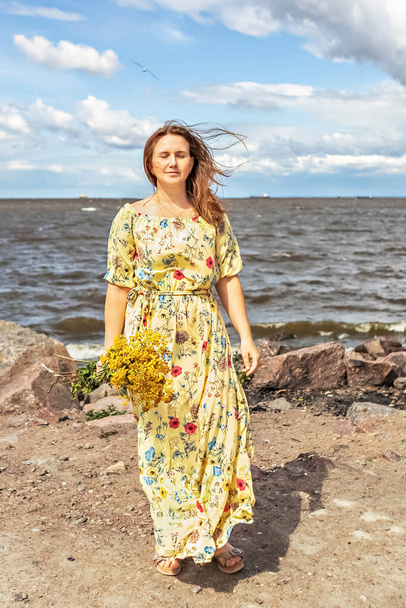 Молодая женщина в жёлтом платье с букетом лакомых кусочков стоит на пляже на ветру. Легкий и воздушный. - Фото, изображение