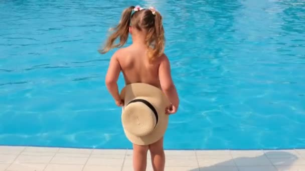 divertida niña de 3 años baila y se esconde detrás de un sombrero de sol en el fondo de agua azul de la piscina o el mar, vista trasera. El concepto de vacaciones de verano, protectores solares, viajes. enfoque selectivo en - Metraje, vídeo