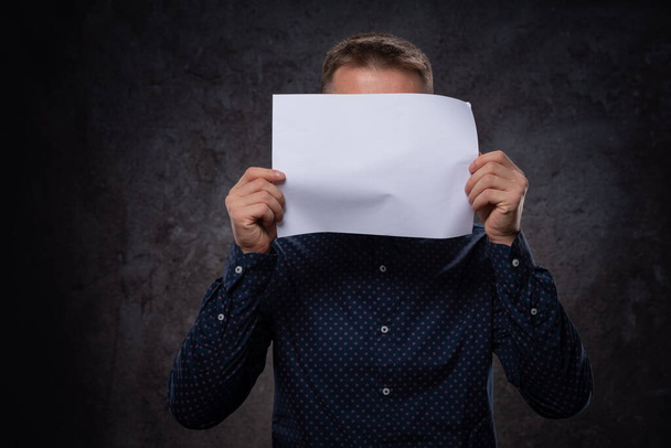 A young European guy holds a blank white sheet of paper in his hands covering his face. For advertising on a dark background - 写真・画像