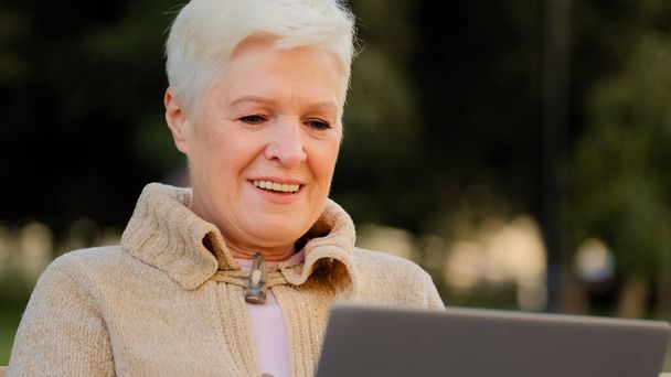 コンピュータのブラウジングやサーフィンインターネットを使用して退職年齢の高齢女性を笑顔ノートパソコンで働く幸せな高齢女性,ニュースをオンラインで読む, PCで興奮した高齢女性のテキストメッセージ屋外に座って - 写真・画像