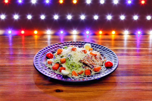 Ensalada César en un plato de barro oscuro sobre una mesa de madera con luces multicolores en el fondo. - Foto, Imagen