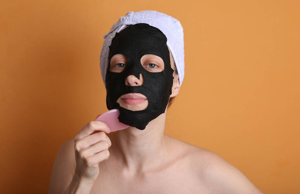 Schöne Frau mit reinigender schwarzer Maske auf dem Gesicht und Handtuch auf dem Kopf. Mädchen massieren Gesicht gua sha Stein. Orange Hintergrund. Gesichtspflege  - Foto, Bild