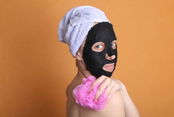 Bella donna in una maschera nera detergente con un asciugamano sulla testa si lava la schiena con un panno su uno sfondo arancione - Foto, immagini