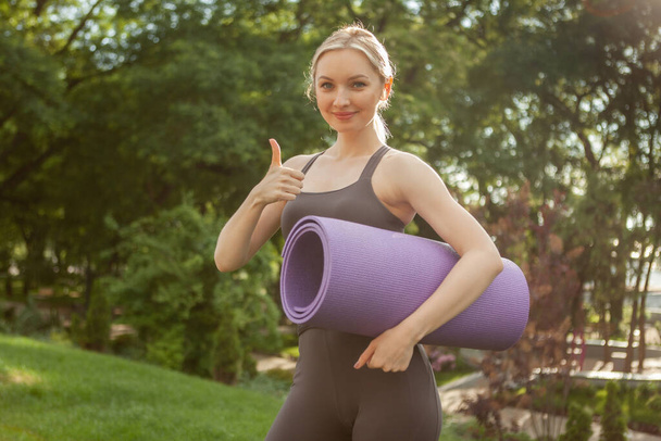 Молодая блондинка держит коврик для йоги и показывает большой палец в парке. Утренняя тренировка. Здоровый образ жизни - Фото, изображение
