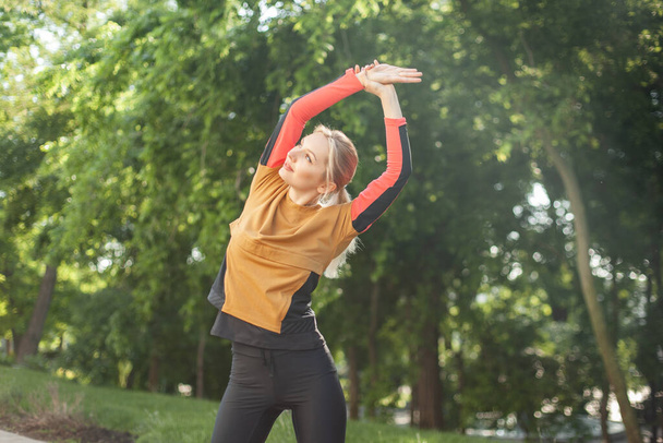 Young fit woman in sportswear practicing stretching before training in park - Фото, изображение