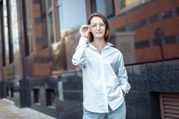 Portrait of a young modern woman in a shirt and glasses on the background of a business building - Photo, Image
