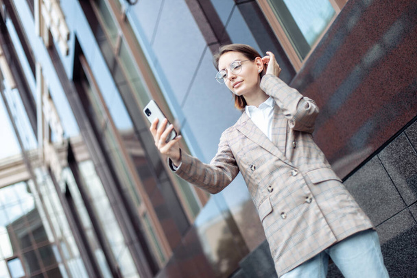 Młoda nowoczesna kobieta biznesu w kurtce i okularach patrząc na ekran smartfona w mieście - Zdjęcie, obraz