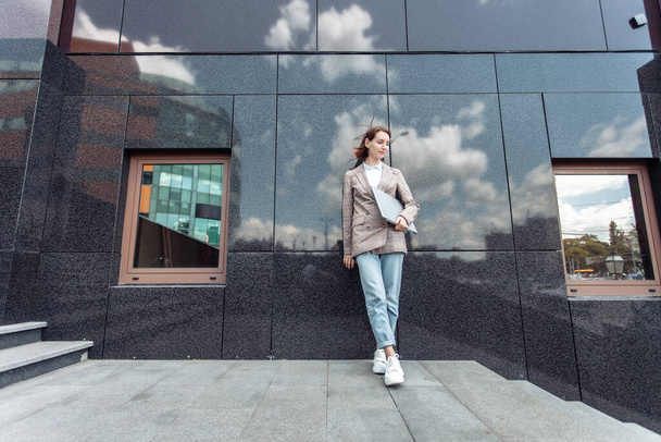 Informelle, junge Geschäftsfrau mit Laptop läuft vor dem Hintergrund eines Geschäftshauses durch die Stadt - Foto, Bild