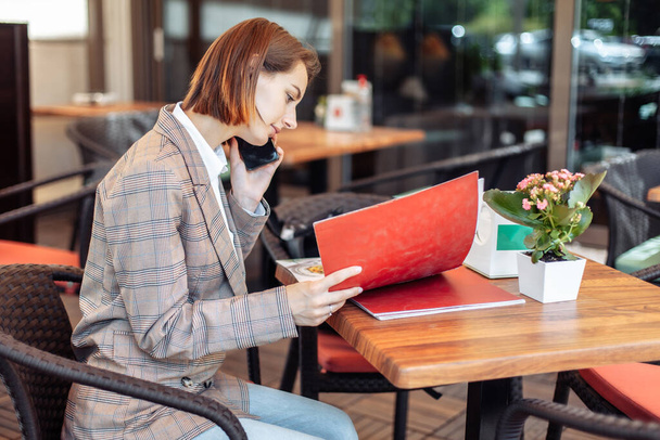 Молодая женщина изучает меню и разговаривает по телефону, сидя за столом в открытом ресторане - Фото, изображение