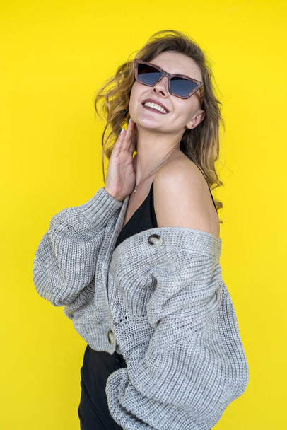 Positive  female model  posing in gray knitted sweater in sunglasses near yellow wall, outdoor - Fotografie, Obrázek