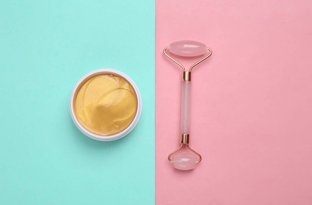 Gold cream jar or patches, facial massage roller on blue pink pastel background. Skin care, lifting face concept - Photo, Image