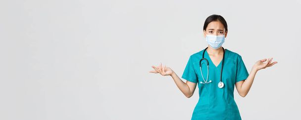 Covid-19, choroba koronawirusowa, koncepcja pracowników służby zdrowia. Dotknięta i zdezorientowana azjatycka pielęgniarka w fartuchu i masce medycznej zadająca pytania, wzruszająca ramionami i wyglądająca na zaniepokojoną, białe tło - Zdjęcie, obraz