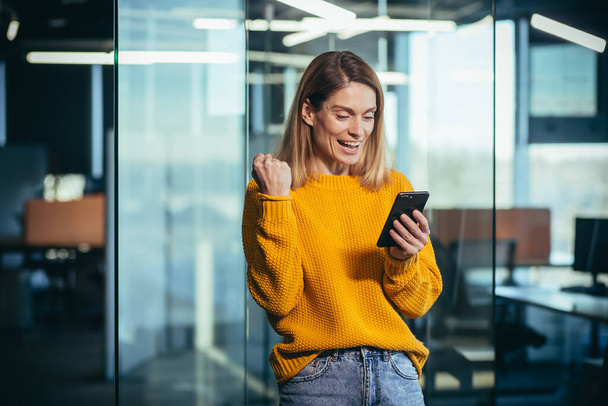 Successful freelancer woman using phone rejoices in winning success, looks at phone, dressed in casual clothes, in modern office, businesswoman boss - Photo, image