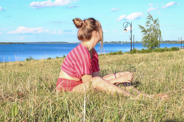 Pembe şortlu ve tişörtlü genç sarışın bir kız çimlerin üzerinde oturuyordu. - Fotoğraf, Görsel