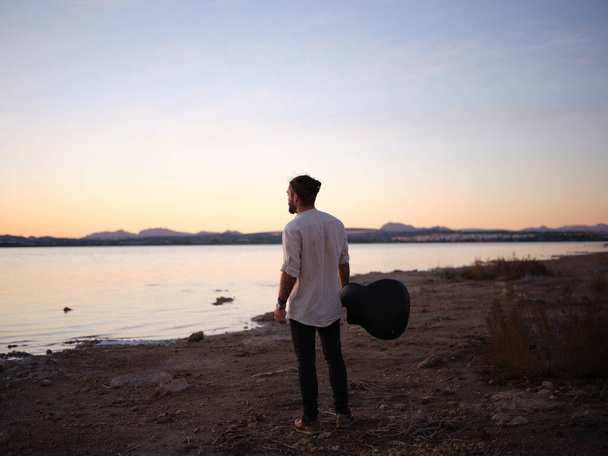 Человек смотрит на горизонт с гитарой перед розовым озером на закате - Фото, изображение