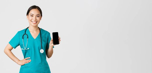 Covid-19, sağlık çalışanları ve online tıp konsepti. Profesyonel bayan Asyalı doktor, önlüklü hemşire akıllı telefon ekranı gösteriyor ve gülümsüyor, internet randevu uygulaması gösteriyor, beyaz arkaplan - Fotoğraf, Görsel
