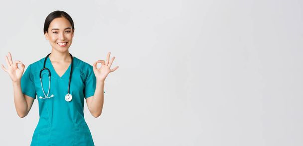 Covid-19, healthcare workers, pandemic concept. Professional confident asian female doctor, intern in scrubs assure patients everything good, showing okay gesture satisfied, smiling pleased - Photo, Image