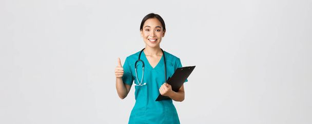 Healthcare workers, preventing virus, quarantine campaign concept. Smiling pretty asian doctor, female intern in scrubs show thumb-up, holding clipboard, bring positive results, white background - Φωτογραφία, εικόνα