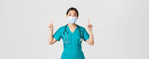 Covid-19, coronavirus disease, healthcare workers concept. Intrigued asian female doctor, physician or intern in medical mask and scrubs, looking curious, pointing fingers up, white background - Photo, Image