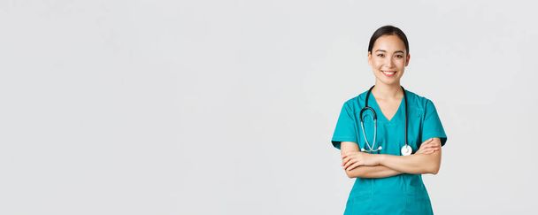 Covid-19, gezondheidswerkers, pandemisch concept. Portret van zelfverzekerde glimlachende, aantrekkelijke Aziatische vrouwelijke verpleegster in scrubs, met stethoscoop, kruisarmen borst en kijken naar camera, witte achtergrond - Foto, afbeelding