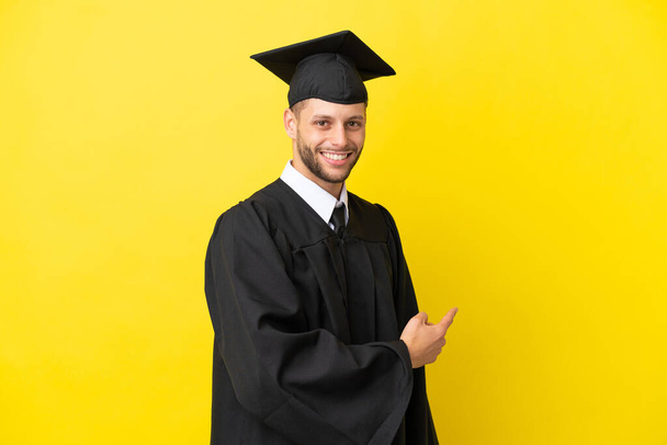 joven universitario graduado caucásico hombre aislado en amarillo fondo apuntando hacia atrás - Foto, Imagen