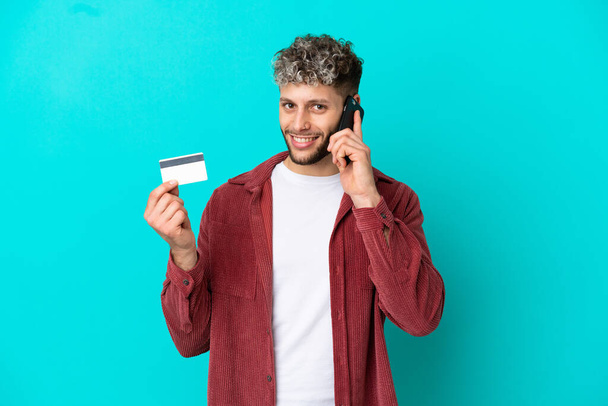 Young handsome caucasian man isolated on blue background keeping a conversation with the mobile phone and holding a credit card - Foto, afbeelding