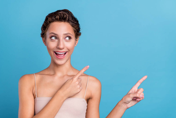 Photo of amazed funky young woman dressed beige top pointing fingers looking empty space smiling isolated blue color background - Foto, afbeelding