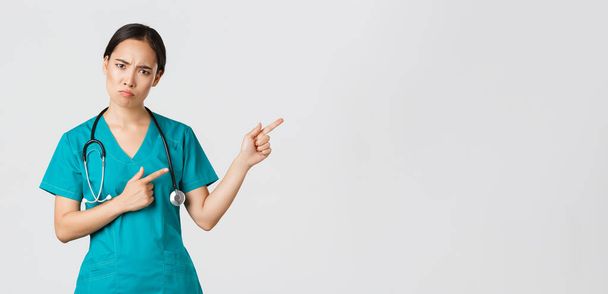 Covid-19, personnel de santé, concept pandémique. Femme thérapeute asiatique déçue et en colère, infirmière en blouse regardant la caméra bouleversée, boudant et pointant vers le coin supérieur droit, explication d'attente - Photo, image