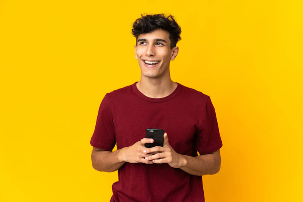 Mladý argentinský muž izolovaný na žlutém pozadí pomocí mobilního telefonu a vzhlíží - Fotografie, Obrázek