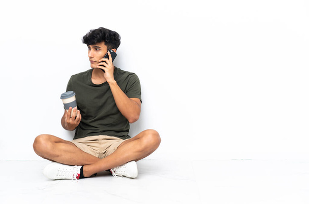 Young Argentinian man sitting on the floor holding coffee to take away and a mobile - 写真・画像