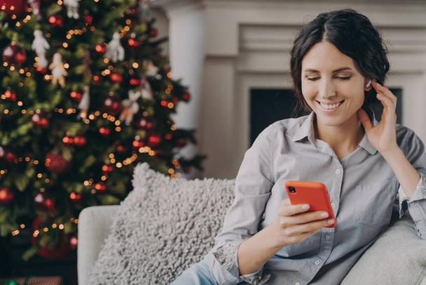 Uvolněný šťastný italský žena sedí v útulné pohovce doma s telefonem během vánočních svátků - Fotografie, Obrázek