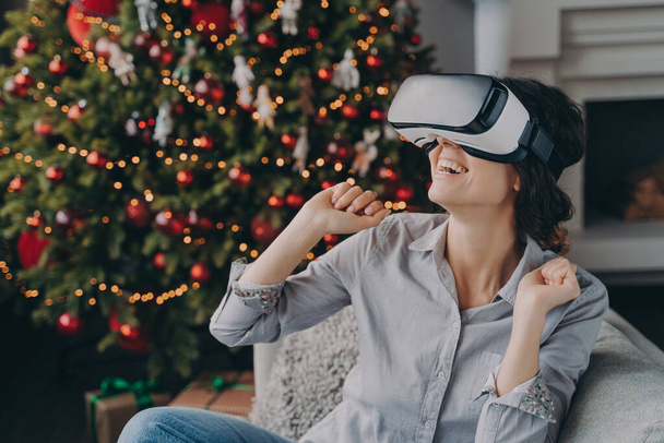 Erstaunte Frau trägt zum ersten Mal Virtual-Reality-Brille, sitzt zu Hause auf Sofa in der Nähe des Weihnachtsbaums - Foto, Bild