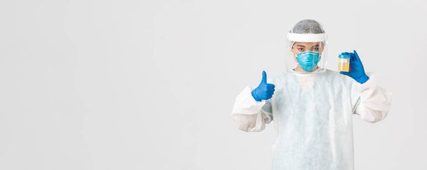 Covid-19, coronavirus disease, healthcare workers concept. Side view of serious-looking tech lab employee, researcher in personal protective equipment examine urine analyzes, show thumbs-up - Photo, Image