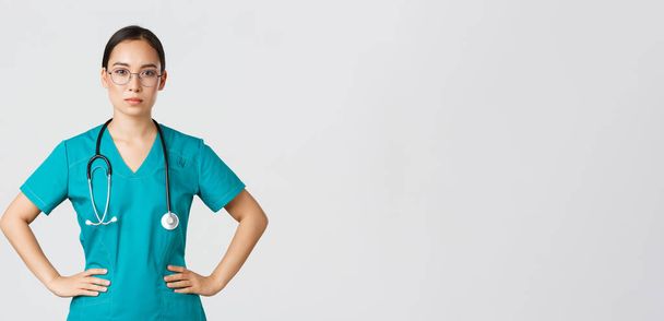 Ковид-19, медицинские работники, пандемия. Уверенная в том, что азиатская медсестра помогает пациентам, работает с коронавирусом, выглядит готовой, носит халаты и очки, белый фон - Фото, изображение