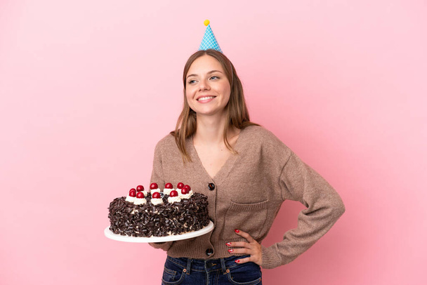 Young Lithuanian woman holding birthday cake isolated on pink background posing with arms at hip and smiling - Photo, Image