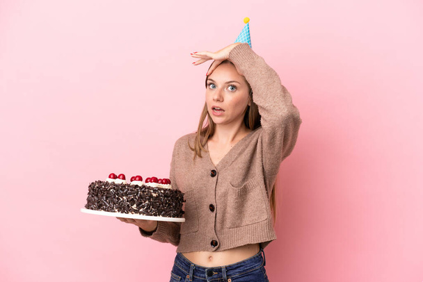 Young Lithuanian woman holding birthday cake isolated on pink background doing surprise gesture while looking to the side - Foto, afbeelding