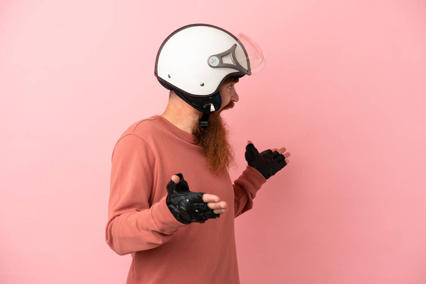 Joven hombre caucásico rojizo con un casco de motocicleta aislado sobre fondo rosa con expresión sorpresa mientras mira al lado - Foto, Imagen