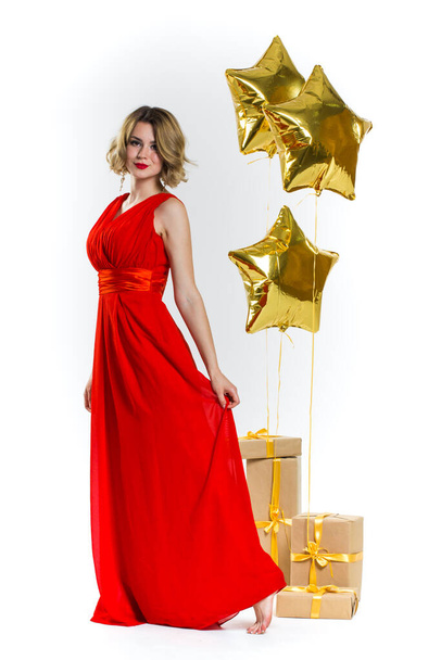 Party photo of elegance sexy lady in red dress with red lips and blond beautiful curly hair, smiling. Background of gold air balloons and gifts. Concept of the sale - Fotó, kép