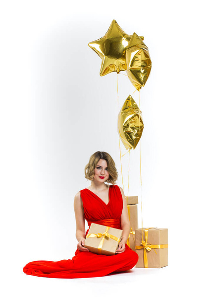 Party photo of elegance sexy lady in red dress with red lips and blond beautiful curly hair, smiling. Background of gold air balloons and gifts. Concept of the sale - Fotó, kép